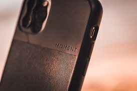A close-up view of a smartphone case with the word 'MOMENT' embossed on it. The case is made of a textured material with a sleek design, featuring a visible camera cutout and side buttons.
