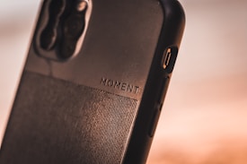 A close-up view of a smartphone case with the word 'MOMENT' embossed on it. The case is made of a textured material with a sleek design, featuring a visible camera cutout and side buttons.
