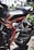 A close-up view of a motorcycle's rear section displaying intricate mechanical parts, a black seat, and a shiny red frame. The background features an ornate metal fence and greenery.