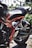 A close-up view of a motorcycle's rear section displaying intricate mechanical parts, a black seat, and a shiny red frame. The background features an ornate metal fence and greenery.