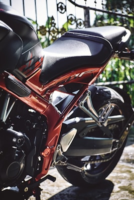 A close-up view of a motorcycle's rear section displaying intricate mechanical parts, a black seat, and a shiny red frame. The background features an ornate metal fence and greenery.