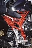 A close-up view of a motorcycle showcasing the intricate details of its frame and suspension. The image highlights a combination of bright red and black components, with visible metallic parts and a textured seat. The design features sharp angles and a modern aesthetic.