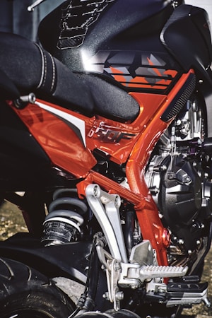 A close-up view of a motorcycle showcasing the intricate details of its frame and suspension. The image highlights a combination of bright red and black components, with visible metallic parts and a textured seat. The design features sharp angles and a modern aesthetic.