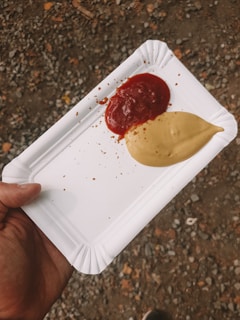 A hand holds a white disposable paper plate with two sauces, one red and one beige, placed side by side. The background consists of a rough, gravel-covered ground.