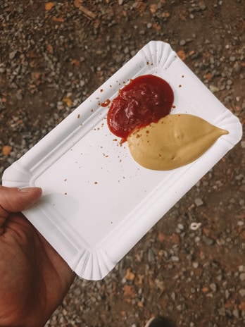 A hand holds a white disposable paper plate with two sauces, one red and one beige, placed side by side. The background consists of a rough, gravel-covered ground.