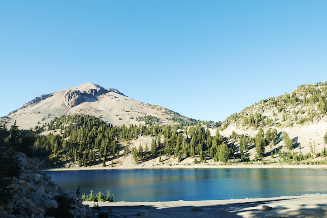 A serene mountain landscape with a clear blue sky and a tranquil lake reflecting the greenery of pine trees and rocky slopes. The mountain is partly covered in trees, with rocky outcrops visible near the peak.