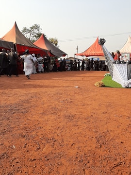 Several people are gathered under large colorful tents on a red sandy ground. The tents are orange and beige with some people dressed in black and white traditional attire. A decorated structure with black, white, and floral patterns is to the right. The scene appears to take place outdoors, with clear skies and trees in the background.
