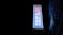 A vertical neon sign with white and red Chinese characters against a blue background is mounted on the side of a building at night. The surrounding area is dark, emphasizing the bright glow of the sign. The building facade features horizontal lines and is partially visible in the dim light.