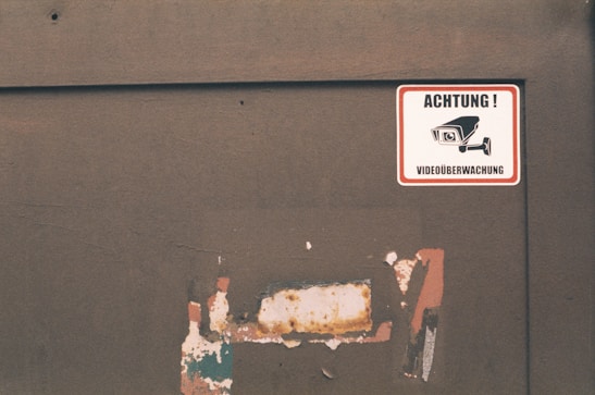 A worn-out wall displays a warning sign in German with the words 'ACHTUNG! VIDEOÜBERWACHUNG,' illustrated with an image of a surveillance camera. The background shows peeling paint and a rusty surface.