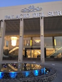 A modern glass-fronted building labeled 'Palacio del Sur' with grand chandeliers visible inside. The facade includes a reflective pool with colorful lights and stone edging, framed by a set of columns.