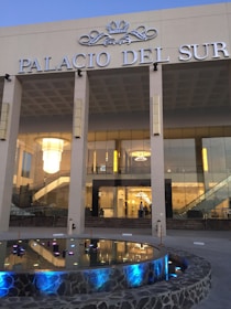 A modern glass-fronted building labeled 'Palacio del Sur' with grand chandeliers visible inside. The facade includes a reflective pool with colorful lights and stone edging, framed by a set of columns.