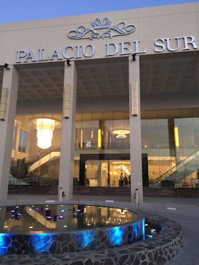 A modern glass-fronted building labeled 'Palacio del Sur' with grand chandeliers visible inside. The facade includes a reflective pool with colorful lights and stone edging, framed by a set of columns.