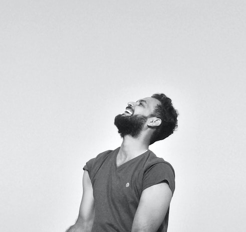 A person with a beard is seen laughing heartily, eyes closed, with their head tilted slightly back. The background is a plain light color, and the person is wearing a short-sleeved t-shirt.