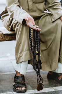 An elderly person wearing traditional clothing is seated, holding a string of prayer beads in their hand. The beads are primarily dark with a few lighter beads interspersed. The person is wearing open-toed sandals and white pants under a long robe.