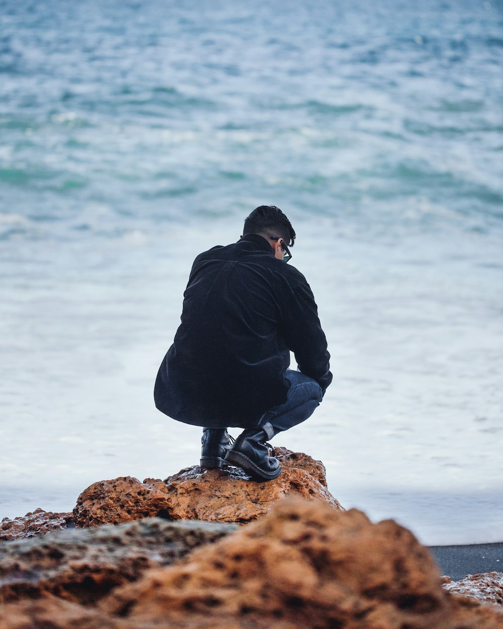 man in black hoodie and black pants sitting on rock formation near body of water during