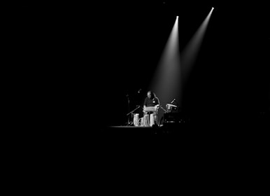 A musician stands in a dimly lit space, focused on playing a set of tablas. The light from two overhead spotlights creates a dramatic effect, highlighting the performer and the instruments amid a stark black background.