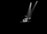 A musician stands in a dimly lit space, focused on playing a set of tablas. The light from two overhead spotlights creates a dramatic effect, highlighting the performer and the instruments amid a stark black background.