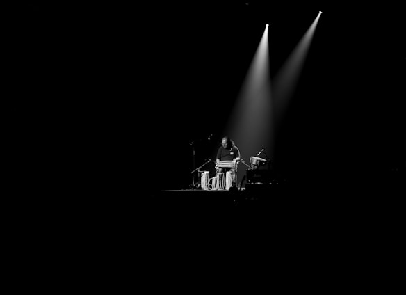 A musician stands in a dimly lit space, focused on playing a set of tablas. The light from two overhead spotlights creates a dramatic effect, highlighting the performer and the instruments amid a stark black background.