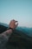A tattooed arm extends into the air, with fingers forming a circular gesture against a backdrop of green hills and distant mountains. The sky is clear with a blue hue.