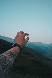 A tattooed arm extends into the air, with fingers forming a circular gesture against a backdrop of green hills and distant mountains. The sky is clear with a blue hue.