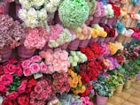 A vibrant display of various colorful flowers arranged neatly in rows of pink baskets. The blooms include roses, carnations, and hydrangeas, showcasing a wide range of colors from pink and red to white, yellow, blue, and purple. The flowers are placed against a tiled floor, creating a lively and cheerful atmosphere.