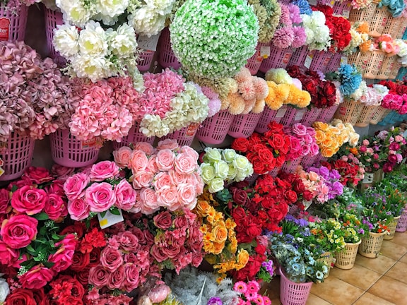 A vibrant display of various colorful flowers arranged neatly in rows of pink baskets. The blooms include roses, carnations, and hydrangeas, showcasing a wide range of colors from pink and red to white, yellow, blue, and purple. The flowers are placed against a tiled floor, creating a lively and cheerful atmosphere.
