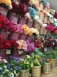A vibrant display of various colorful flowers arranged in plastic baskets. The flowers include roses, daisies, and other varieties, showcasing a wide assortment of colors such as red, pink, yellow, orange, blue, and white. Price tags are attached to some of the baskets, indicating that the flowers are for sale.