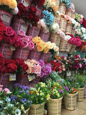 A vibrant display of various colorful flowers arranged in plastic baskets. The flowers include roses, daisies, and other varieties, showcasing a wide assortment of colors such as red, pink, yellow, orange, blue, and white. Price tags are attached to some of the baskets, indicating that the flowers are for sale.