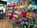 A vibrant flower shop displays an array of artificial flowers organized in baskets on the walls. The flowers, showcasing a multitude of colors including red, pink, orange, and white, provide a visually appealing scene. The shop interior features tiled flooring and a partial view of green foliage spheres.