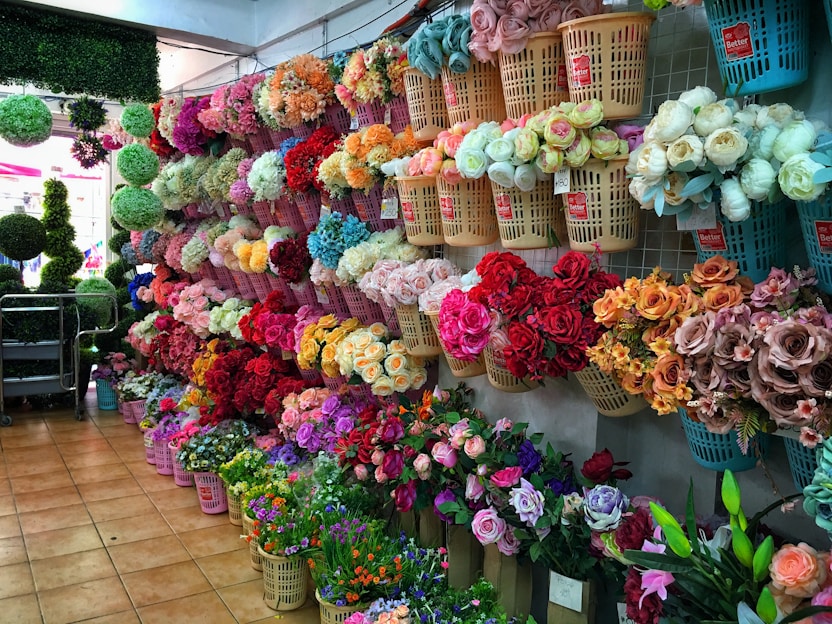 A vibrant flower shop displays an array of artificial flowers organized in baskets on the walls. The flowers, showcasing a multitude of colors including red, pink, orange, and white, provide a visually appealing scene. The shop interior features tiled flooring and a partial view of green foliage spheres.