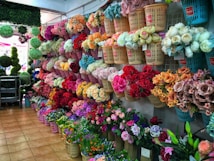 A vibrant flower shop displays an array of artificial flowers organized in baskets on the walls. The flowers, showcasing a multitude of colors including red, pink, orange, and white, provide a visually appealing scene. The shop interior features tiled flooring and a partial view of green foliage spheres.