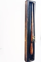 A wooden object resembling an old musket is encased in a narrow, dark wooden shadow box. The object has a long metal rod attached to it.