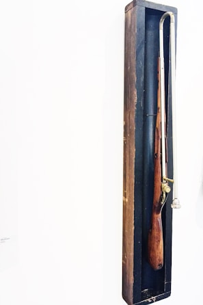 A wooden object resembling an old musket is encased in a narrow, dark wooden shadow box. The object has a long metal rod attached to it.