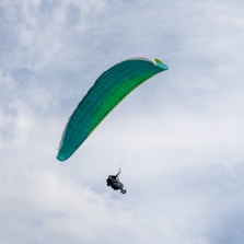 A person is paragliding against a backdrop of a partly cloudy sky. The parachute is a vibrant green color, and the person appears to be securely strapped in, enjoying a flight through the sky.