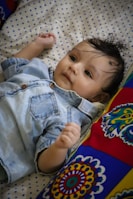 A baby wearing a denim shirt lies on a polka dot patterned surface, surrounded by a colorful patterned pillow. The baby seems content and relaxed, with wisps of hair falling on the forehead.