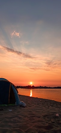 A serene beach scene at sunset with crystal-clear water and a cozy glamping tent nearby.