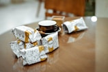 A wooden table with several packaged snacks featuring doodle-style art and the brand name 'Biscoman'. There is a black coffee cup with a white interior resting on the table. The background appears to be a cozy indoor setting with blurred elements.