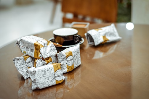 A wooden table with several packaged snacks featuring doodle-style art and the brand name 'Biscoman'. There is a black coffee cup with a white interior resting on the table. The background appears to be a cozy indoor setting with blurred elements.