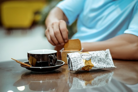 A person wearing a light blue shirt is reaching for a biscuit from a packaging labeled 'Bisconama.' A black coffee cup with a saucer and a wooden stirrer rests on a reflective surface.