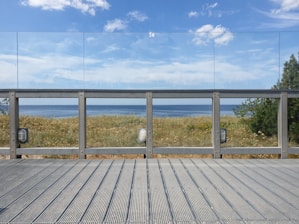 A wooden deck leads to a glass railing overlooking a scenic view of the ocean. Behind the railing, dried grasses and wildflowers cover the ground, with a few trees visible on the right. A clear blue sky with scattered clouds adds to the tranquil setting.