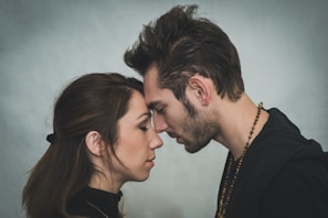 A man and woman stand facing each other with their foreheads gently touching, eyes closed, suggesting a moment of intimacy and connection. Both individuals have relaxed expressions, with the man's hand resting on the back of the woman's head.