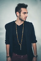 A man with short, styled hair is standing sideways, looking to his right. He is wearing a black V-neck shirt underneath a dark jacket, and a long beaded necklace. His arms are relaxed by his sides, and he has a watch on his left wrist.
