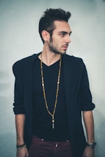 A man with short, styled hair is standing sideways, looking to his right. He is wearing a black V-neck shirt underneath a dark jacket, and a long beaded necklace. His arms are relaxed by his sides, and he has a watch on his left wrist.