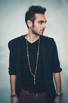 A man with short, styled hair is standing sideways, looking to his right. He is wearing a black V-neck shirt underneath a dark jacket, and a long beaded necklace. His arms are relaxed by his sides, and he has a watch on his left wrist.