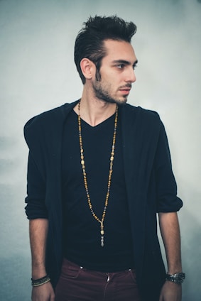 A man with short, styled hair is standing sideways, looking to his right. He is wearing a black V-neck shirt underneath a dark jacket, and a long beaded necklace. His arms are relaxed by his sides, and he has a watch on his left wrist.