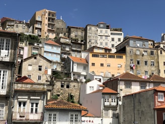 A densely packed hillside neighborhood with a variety of old residential buildings. Houses are stacked closely together, featuring colorful exteriors, with shades of blue, pink, and beige. Some buildings display signs of wear, with peeling paint and rustic stone structures. Red-tiled roofs add a traditional touch, and there are a few satellite dishes and clotheslines visible.
