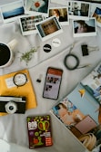 A collection of personal items and photographs is arranged on a light-colored surface. There are several instant photos depicting various scenes including people, interiors, and landscapes. A vintage camera and a smartphone displaying a webpage are present alongside a packet of snacks labeled 'BOB Snail.' A cup of coffee, a small plant sprig, and a few stationery items, such as binder clips and a cable, add to the assortment. A yellow envelope with handwritten text is partially covered by the camera, and an open magazine featuring an interior design is also visible.