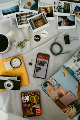 A collection of personal items and photographs is arranged on a light-colored surface. There are several instant photos depicting various scenes including people, interiors, and landscapes. A vintage camera and a smartphone displaying a webpage are present alongside a packet of snacks labeled 'BOB Snail.' A cup of coffee, a small plant sprig, and a few stationery items, such as binder clips and a cable, add to the assortment. A yellow envelope with handwritten text is partially covered by the camera, and an open magazine featuring an interior design is also visible.