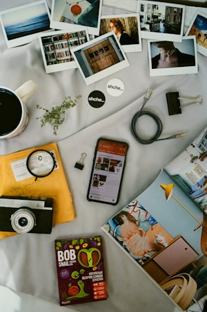 A collection of personal items and photographs is arranged on a light-colored surface. There are several instant photos depicting various scenes including people, interiors, and landscapes. A vintage camera and a smartphone displaying a webpage are present alongside a packet of snacks labeled 'BOB Snail.' A cup of coffee, a small plant sprig, and a few stationery items, such as binder clips and a cable, add to the assortment. A yellow envelope with handwritten text is partially covered by the camera, and an open magazine featuring an interior design is also visible.