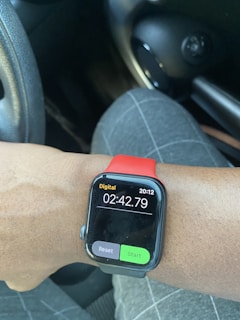 A smartwatch with a red band is displaying a digital timer reading 02:42.79. The watch is on the wrist of a person wearing plaid-patterned pants. The background shows part of a vehicle's interior, including a steering wheel and a round air vent.